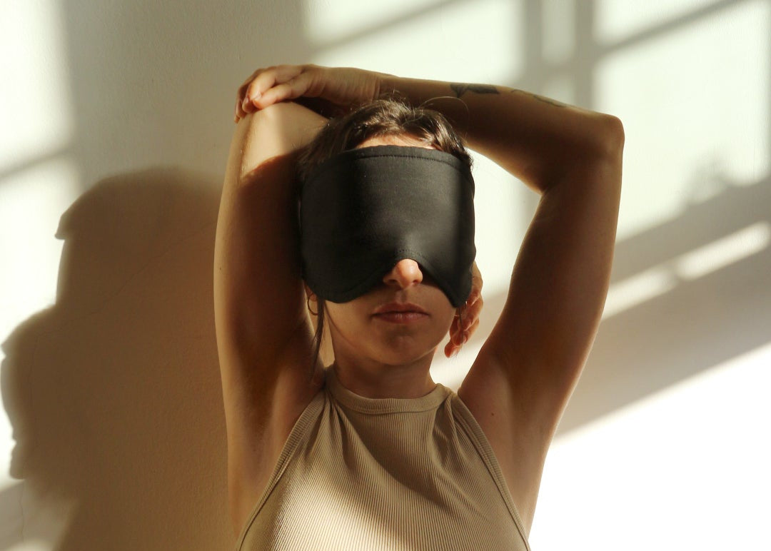 Person sitting on a bed with arms raised, wearing a black mask and holding a candle.